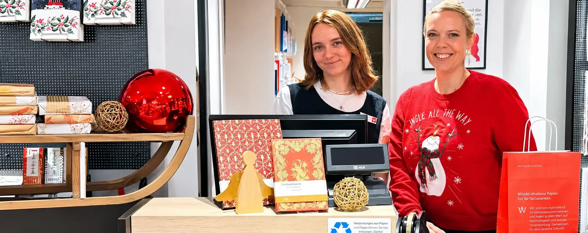 Zwei Heymann-Kolleginnen an der weihnachtlich dekorierten Kasse mit Geschenkpapier und Weihnachtskarten.